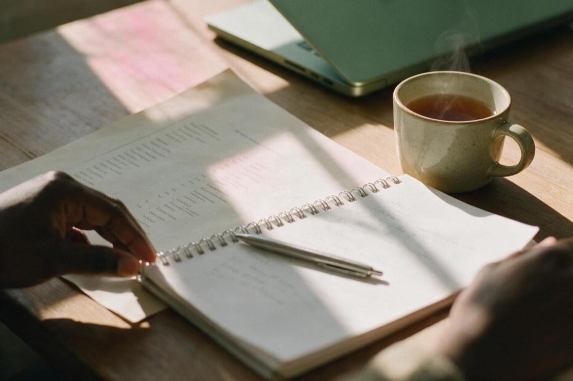 A desk in morning light with a printed staff list, an open notebook, a pen and a cup of tea.